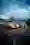 Two rally cars are parked on a road at dusk. In the foreground is a bright yellow-coloured Volvo PV444 or PV544 with start number 05 and its headlights switched on. Directly behind it is a white, red and blue car, possibly a Lancia, also with its headlights switched on.